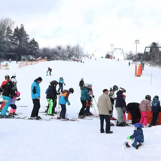 Eine Gruppe von Menschen, einschließlich Kindern, Ski auf einem verschneiten Berg. Einige stehen, andere sitzen. Bäume und eine Skilift im Hintergrund.