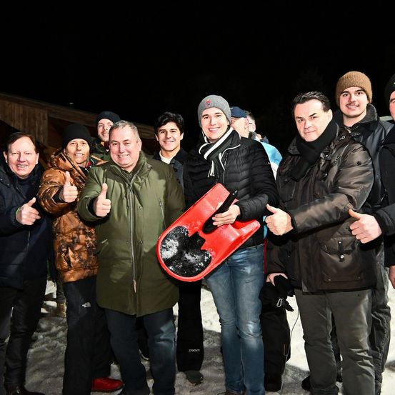 Eine Gruppe von Männern in Winterkleidung steht im Schnee, hält einen roten Schlitten, lächelt und gibt Daumen hoch. Hinter ihnen steht ein Gebäude mit einem dunklen Himmel im Hintergrund.