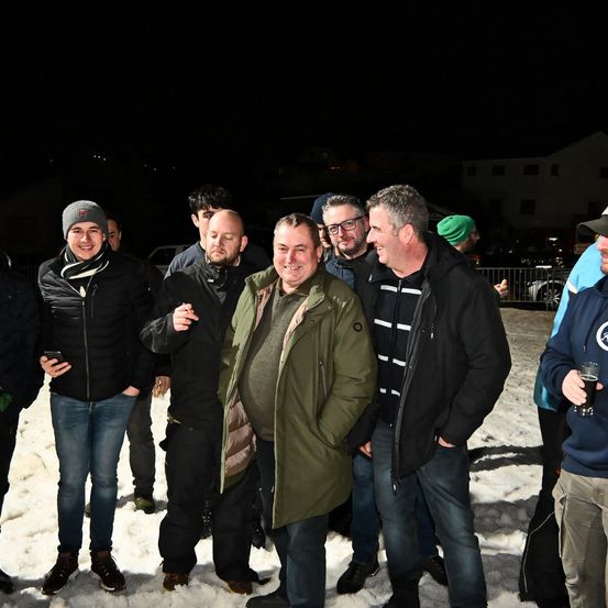 Eine Gruppe von Männern in Winterkleidung steht nachts im Schnee und posiert für ein Foto. Ein Mann hält ein Glas Bier. Im Hintergrund ist ein Haus und ein Auto zu sehen.