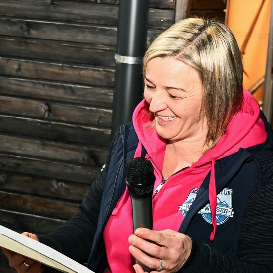 Eine Frau mit blondem Haar trägt eine pinke Kapuzenjacke und eine schwarze Jacke. Sie hält ein Mikrofon und ein Stück Papier. Sie lächelt und scheint aus dem Papier zu lesen. Hinter ihr befindet sich eine Holzwand.