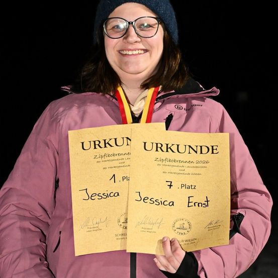Eine lächelnde Frau in einer rosa Jacke hält zwei gelbe Urkunden, eine mit 'Jessica 1. Platz' und die andere mit 'Jessica 7. Platz'. Sie trägt eine Mütze, eine Brille und eine Medaille. Der Hintergrund ist dunkel.