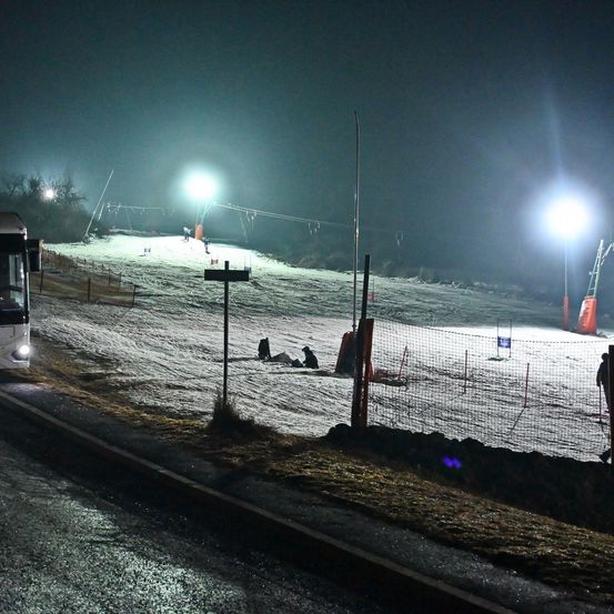 Nachtansicht einer Skipiste mit zwei liegenden Personen. Helle Lichter beleuchten den Bereich, ein Bus und ein Zaun sind im Vordergrund sichtbar.