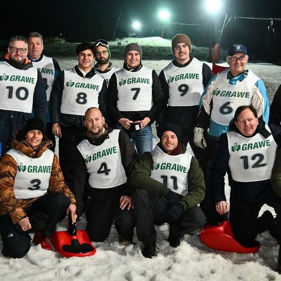 Eine Gruppe von Personen in Winterkleidung posiert nachts für ein Foto, alle tragen nummerierte Westen. Sie stehen und sitzen auf Schnee mit Schaufeln vor ihnen.