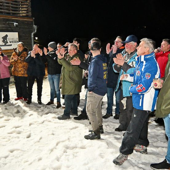 Eine Gruppe von Menschen steht bei Nacht im Schnee und klatscht und jubelt. Sie tragen Winterkleidung. Ein Banner mit Text befindet sich an der Wand hinter ihnen.