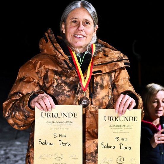 Eine Frau hält zwei Urkunden und eine Medaille. Die Urkunden sind mit '3. Platz' und '15. Platz' beschriftet, jeweils mit dem Namen 'Sabina Doria'. Die Frau trägt eine Tarnjacke und eine Medaille um den Hals. Hinter ihr hält eine weitere Frau ein Mikrofon.