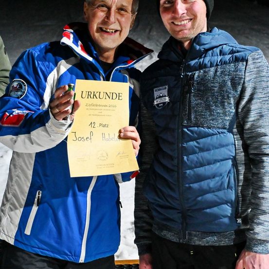 Zwei Männer stehen im Schnee und lächeln. Ein Mann hält ein Zertifikat, das Platz 12 anzeigt. Der Mann auf der linken Seite hält eine Bierdose.