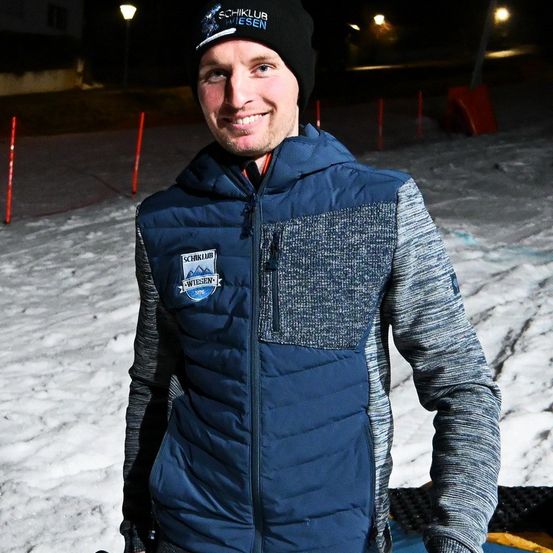 Ein Mann steht bei Nacht auf einem schneebedeckten Feld, trägt eine dunkelblaue Jacke mit Logo und eine schwarze Mütze. Der Boden ist schneebedeckt und hat rote Pfosten und Lichter.