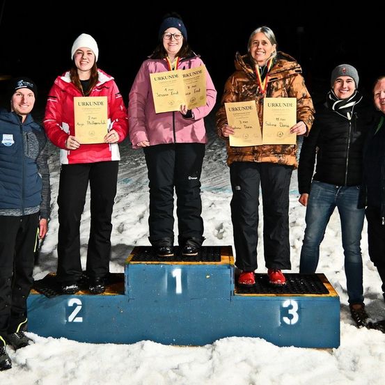 Fünf Personen stehen auf einem verschneiten Podest, wobei die Drittplatzierte rechts steht. Die Erstplatzierte hält ein Zertifikat, ebenso wie die Zweit- und Drittplatzierte.