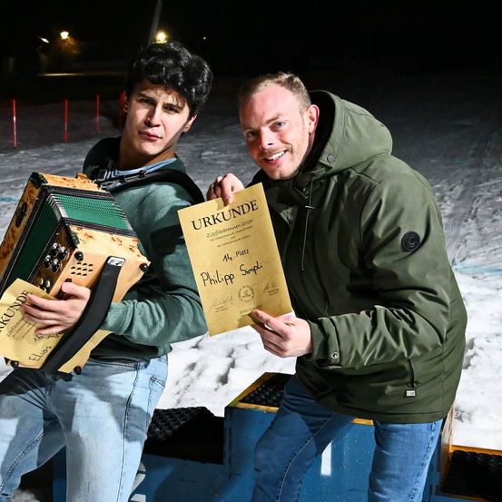 Zwei Männer posieren nachts für ein Foto. Der Mann auf der linken Seite hält ein Akkordeon und der Mann auf der rechten Seite hält ein Zertifikat. Beide tragen grüne Jacken und Jeans. Im Hintergrund ist Schnee zu sehen.
