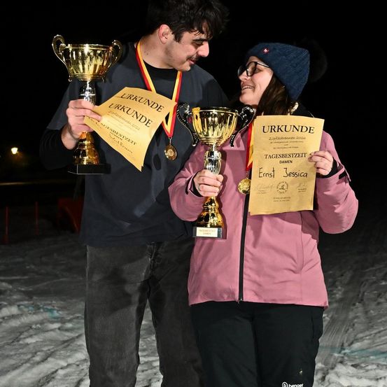 Ein Mann und eine Frau stehen im Schnee, lächelnd und posierend mit goldenen Trophäen und Urkunden. Der Mann hält eine Urkunde mit dem Titel 'Urkunde' und die Frau hält eine Urkunde mit dem Titel 'Tagesbestzeit Damen'.