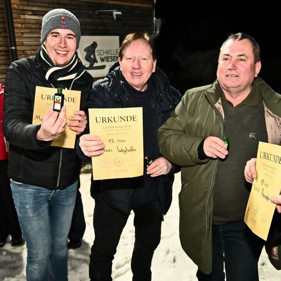 Drei Männer stehen im Schnee und lächeln. Der Mann auf der linken Seite hält eine kleine Flasche und ein Zertifikat. Der Mann in der Mitte hält ein größeres Zertifikat. Der Mann auf der rechten Seite hält eine Flasche und ein Zertifikat. Hinter ihnen ist eine Holzwand mit einem Schild.