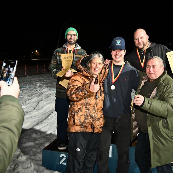 Eine Gruppe von Menschen steht nachts auf einem Podest im Schnee. Sie alle haben Medaillen um den Hals. Ein Mann hält ein Schild und lächelt, während ein anderer eine Flasche hält. Eine Person macht ein Foto von ihnen mit einem Handy.