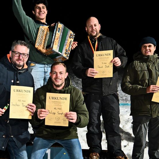 Fünf Männer stehen im Schnee und halten Urkunden. Einer spielt Akkordeon. Ein anderer hält ein Bier. Ein weiterer trägt eine Medaille. Sie alle lächeln.