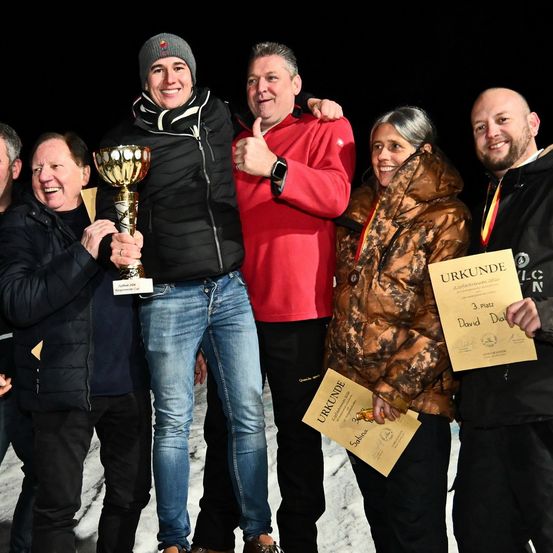 Eine Gruppe von Personen, in warmer Kleidung gekleidet, feiert mit Pokalen und Urkunden auf einer verschneiten Oberfläche. Der Mann in der Mitte hält einen goldenen Pokal, während andere Daumen hochhalten.