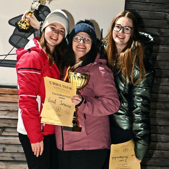 Drei Frauen in Winterkleidung posieren für ein Foto mit Trophäen. Eine hält ein Zertifikat für Ernst Jessica, während die anderen lächeln und in die Kamera schauen.
