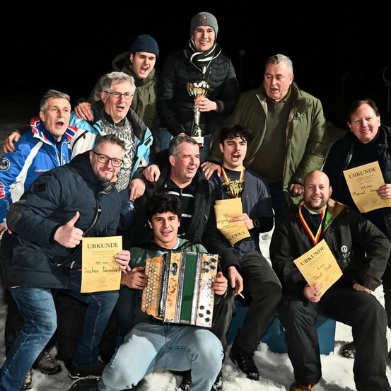 Eine Gruppe von Männern posiert im Schnee für ein Foto, hält Auszeichnungen und einen Pokal. Ein Mann in der Mitte spielt Akkordeon, während andere jubeln.