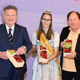 Eine gekrönte Frau steht zwischen zwei Männern, alle halten Schachteln mit Erdbeeren vor einem lila Stadt Wien-Banner.