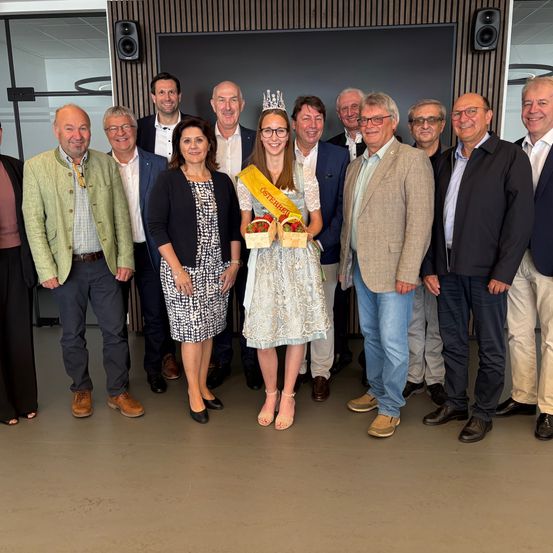 Eine Gruppe von Erwachsenen posiert für ein Foto, darunter eine Frau, die eine gelbe Schärpe und einen Korb mit Erdbeeren hält. Sie alle lächeln und stehen zusammen.