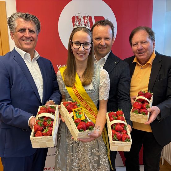 Fünf Personen, drei Männer und zwei Frauen, stehen vor einem roten Hintergrund und lächeln. Die Frau in der Mitte trägt eine Krone und hält eine Box mit Erdbeeren. Die Männer tragen Anzüge, und einer hält eine Box mit Erdbeeren.
