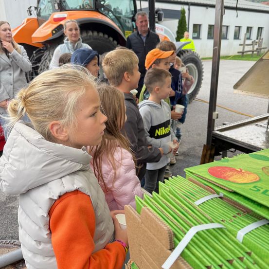 Eine Gruppe von Kindern und Erwachsenen steht in einer Reihe, wahrscheinlich in der Schlange, um etwas zu erhalten. Sie halten Tassen. Ein großer Traktor steht hinter ihnen.