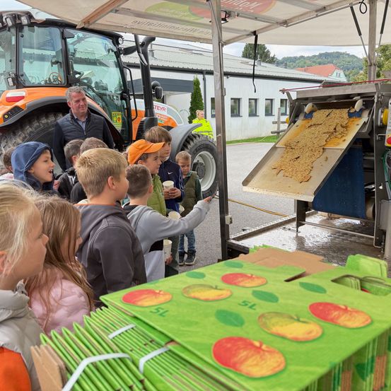 Eine Gruppe von Kindern beobachtet einen Mann, der eine Maschine auf einem Bauernhof bedient. Die Maschine verarbeitet Äpfel, und Kisten mit Äpfeln sind sichtbar.