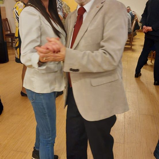 Ein Mann und eine Frau tanzen bei einer Indoor-Veranstaltung zusammen. Der Mann trägt Anzug und Krawatte, während die Frau einen weißen Blazer und Jeans trägt.