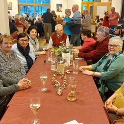 Eine Gruppe von Menschen, meist älterer, sitzt an einem langen Tisch in einem Raum. Einige tragen Brillen und lächeln. Der Tisch hat Weingläser, eine Blumenarrangement und andere Gegenstände.