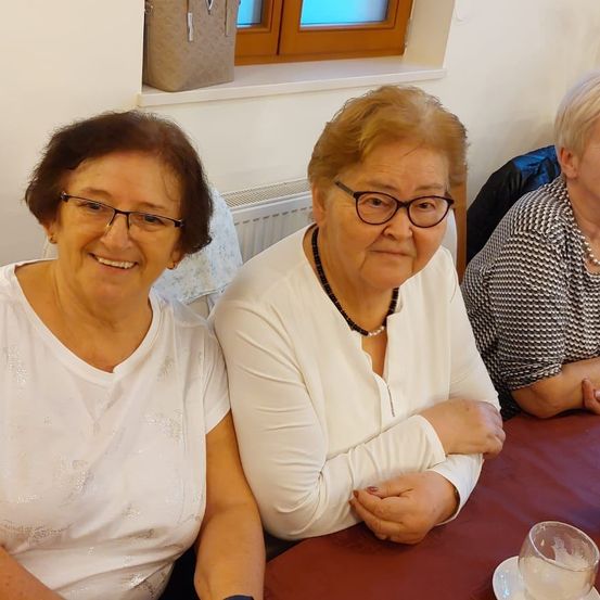 Drei ältere Frauen sitzen an einem Tisch und lächeln. Die Frau in der Mitte trägt ein weißes Oberteil und eine Halskette. Die Frau auf der linken Seite trägt eine Brille und eine Uhr. Hinter ihnen ist ein Fenster mit einer Tasche auf der Fensterbank.