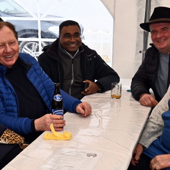Drei Männer sitzen an einem Tisch, zwei lächeln, einer hält eine Bierflasche. Eine vierte Person in einem Leopardenmuster-Jacke sitzt neben ihnen. Dahinter ein Autofenster mit einer Reflexion.