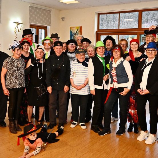 Eine Gruppe von Menschen in Kostümen und Hüten posiert für ein Foto in einem Raum mit Holzböden und großen Fenstern.