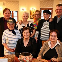 Eine Gruppe von Menschen posiert für ein Foto in einem Raum mit weißen Wänden, Fenstern und Körben auf dem Tisch. Einige tragen Brillen und Halsketten.