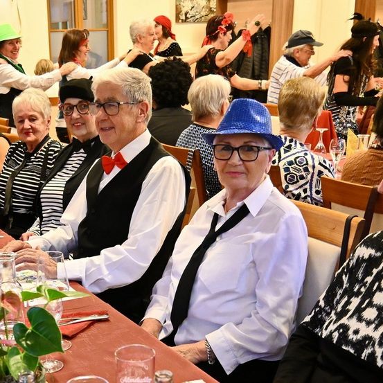 Eine Gruppe älterer Menschen, gekleidet in festlicher Kleidung, sitzt an einem Tisch, möglicherweise bei einem Treffen oder einer Veranstaltung. Sie tragen Hüte, Brillen und Krawatten. Einige haben Blumen auf dem Kopf. Der Tisch ist mit Gläsern und einer Pflanze gedeckt.