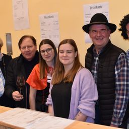 Eine Gruppe von Menschen steht lächelnd für ein Foto hinter einer Theke. Ein Mann hält ein Weinglas, und eine Speisekarte ist auf der Theke sichtbar. Dahinter hängen Weinkarten an der Wand.