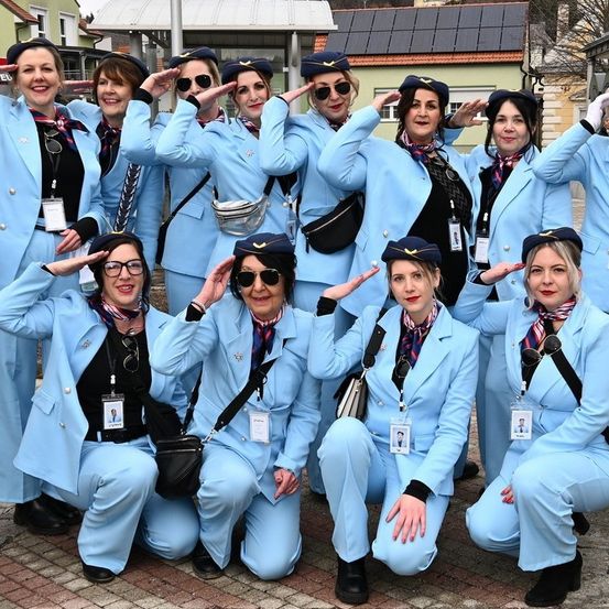 Eine Gruppe von Frauen in passenden hellblauen Anzügen posiert für ein Foto im Freien, alle tragen Hüte und Sonnenbrillen. Sie lächeln und machen eine Salutgeste.