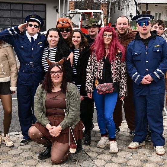 Eine Gruppe von Menschen in verschiedenen Kostümen posiert für ein Foto auf einer gepflasterten Straße. Einige tragen Uniformen, Brillen, Hüte und Schuhe. Im Hintergrund befinden sich Gebäude und ein Fahrzeug.