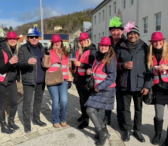 Eine Gruppe von Menschen in rosa Hüten und Warnwesten posiert für ein Foto auf einer Straße. Sie halten Getränke in den Händen, und einige lächeln. Das Wetter ist sonnig.