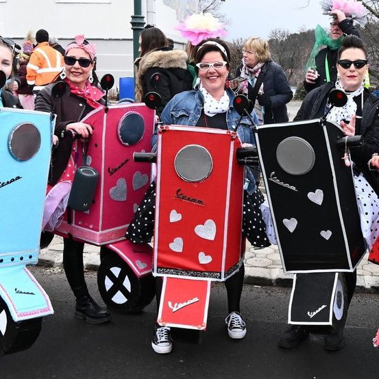 Fünf Frauen in Vespa-Kostümen mit Herzen, die in einer Reihe in einem Umzug stehen. Eine hat ein rosa Kostüm mit einer rosa Schleife, eine andere hat eine Jeansjacke.