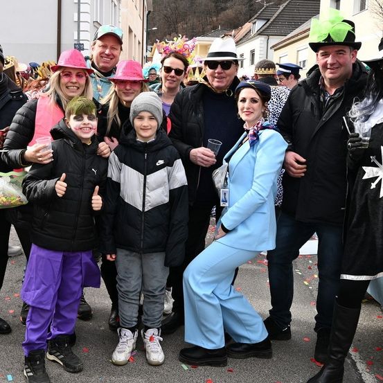 Eine Gruppe von Menschen, einige in Kostümen, posiert für ein Foto auf einer Straße. Sie lächeln, mit Konfetti auf dem Boden.
