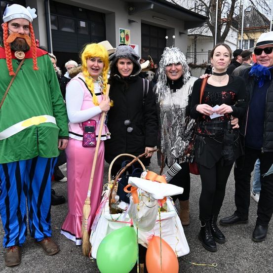 Eine Gruppe von Menschen in verschiedenen Kostümen steht draußen und lächelt für ein Foto. Ein Korb mit einem Kürbis und Luftballons steht vor ihnen.