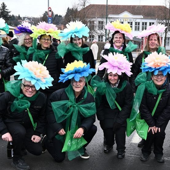 Eine Gruppe von Menschen in Kostümen mit Blumenhüten posiert für ein Foto auf der Straße vor einem Schulgebäude.