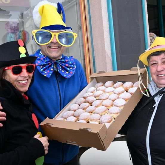 Drei Menschen in Kostümen halten eine Schachtel mit Donuts vor einem Laden. Einer hält die Schachtel, während die anderen lächeln.