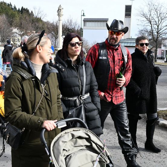 Vier Personen, zwei Frauen und zwei Männer, stehen mit einem Kinderwagen draußen. Die Frauen tragen Wintermäntel, eine trägt eine Sonnenbrille. Die Männer sind in Schwarz und Rot gekleidet, einer hält eine Flasche. Bäume und ein Gebäude sind im Hintergrund.
