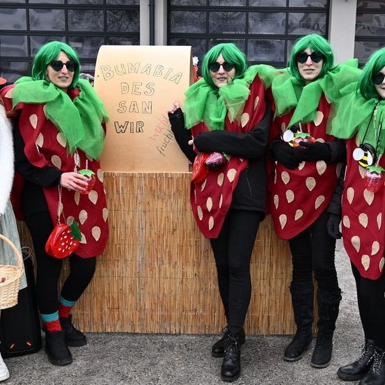 Vier Frauen in Erdbeerkostümen stehen zusammen. Jede trägt eine grüne Perücke, Sonnenbrillen und eine rote Erdbeertasche. Sie stehen vor einem Holzstand mit einem Schild, auf dem 'BUMABIA DES SAN WR' steht. Der Schauplatz scheint ein Outdoor-Event zu sein.