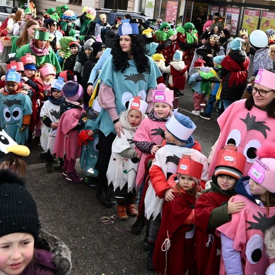 Eine Gruppe von Menschen, darunter Kinder in Kostümen, versammelt sich im Freien. Einige tragen Hüte und Brillen, andere haben Luftballons. Eine Frau mit langen Haaren hält ein Kind in einem Kostüm. Im Hintergrund sind Gebäude und Autos zu sehen.