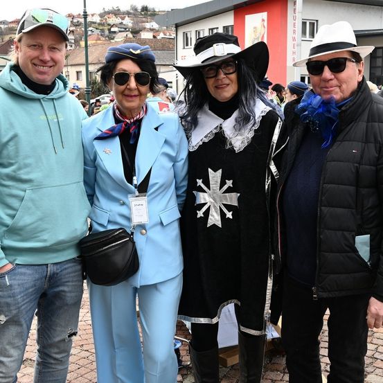 Vier Menschen in formeller Kleidung posieren für ein Foto im Freien. Die Frau in der Mitte trägt einen blauen Anzug, während die Frau rechts eine schwarze Robe mit Silberbesatz trägt. Der Mann rechts trägt eine schwarze Jacke.