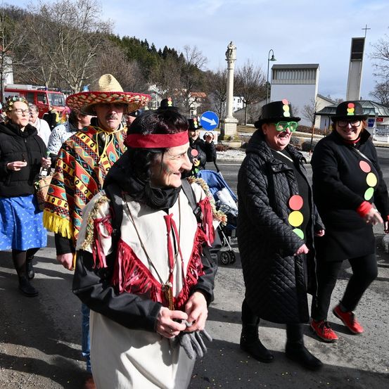 Eine Gruppe von Menschen in festlicher Kleidung geht die Straße entlang. Eine Frau in einem weißen Kleid und einem roten Stirnband führt die Gruppe an. Hinter ihr ein Mann in einem Sombrero und eine Frau, die eine Mütze mit Ampeln trägt. Sie sind von anderen umgeben, darunter eine Person, die einen Kinderwagen schiebt. Bäume und ein Gebäude sind im Hintergrund.