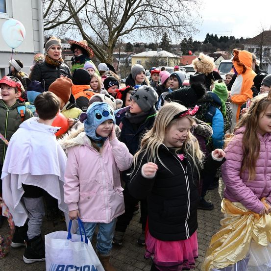 Kinder in Kostümen versammeln sich vor einem Gebäude, eines hält eine Tasche mit der Aufschrift Action, ein anderes hält einen Ballon. Eine Frau mit einem Schal spricht im Hintergrund.