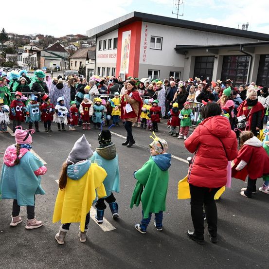Kinder in Kostümen sind bei einer Straßenparade versammelt, Erwachsene beobachten im Hintergrund. Eine Frau in Rot steht vorne. Gebäude mit Schildern sind sichtbar.