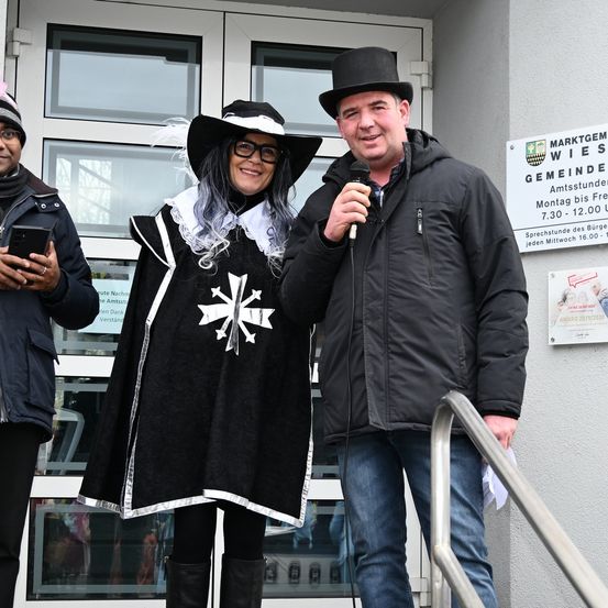 Drei Personen stehen draußen, eine mit einem Mikrofon. Die Frau in Hut und Mantel lächelt. Der Mann im Zylinder hält ein Mikrofon. Hinter ihnen ist eine Wand mit Informationen über eine Veranstaltung.