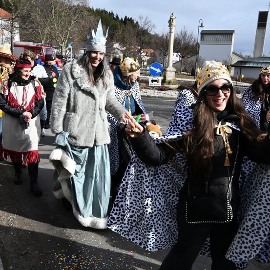 Eine Gruppe von Frauen in festlicher Kleidung geht lächelnd Hand in Hand die Straße entlang. Sie tragen Kronen und bunte Kleider. Dahinter sind weitere Menschen in Kostümen und ein Feuerwehrauto zu sehen.